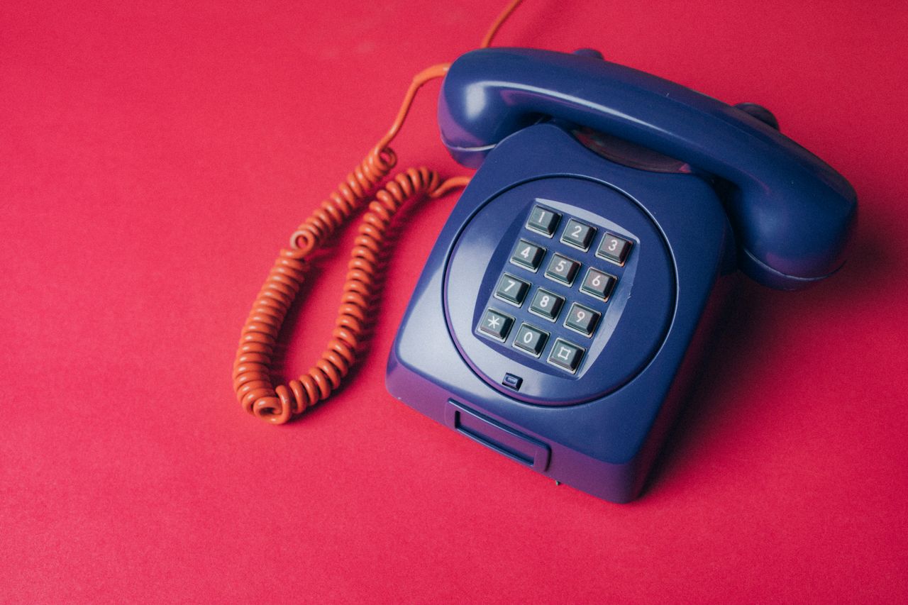 A classic blue rotary phone with a coiled cord on a vibrant red backdrop, exuding nostalgia.